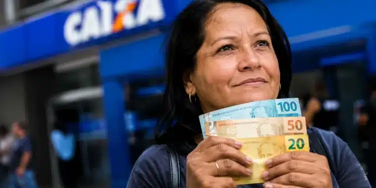 Mulher segura cédulas de real em frente a uma agência da Caixa, representando o período de pagamentos dos benefícios do INSS de até um salário mínimo em agosto.