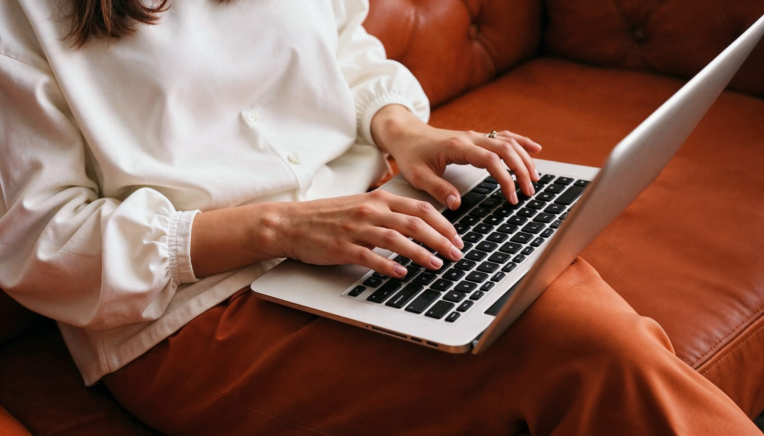 Mulher com blusa branca digitando em notebook, sentada em sofá caramelo, simbolizando a consulta de cursos e instiutições no e-MEC.