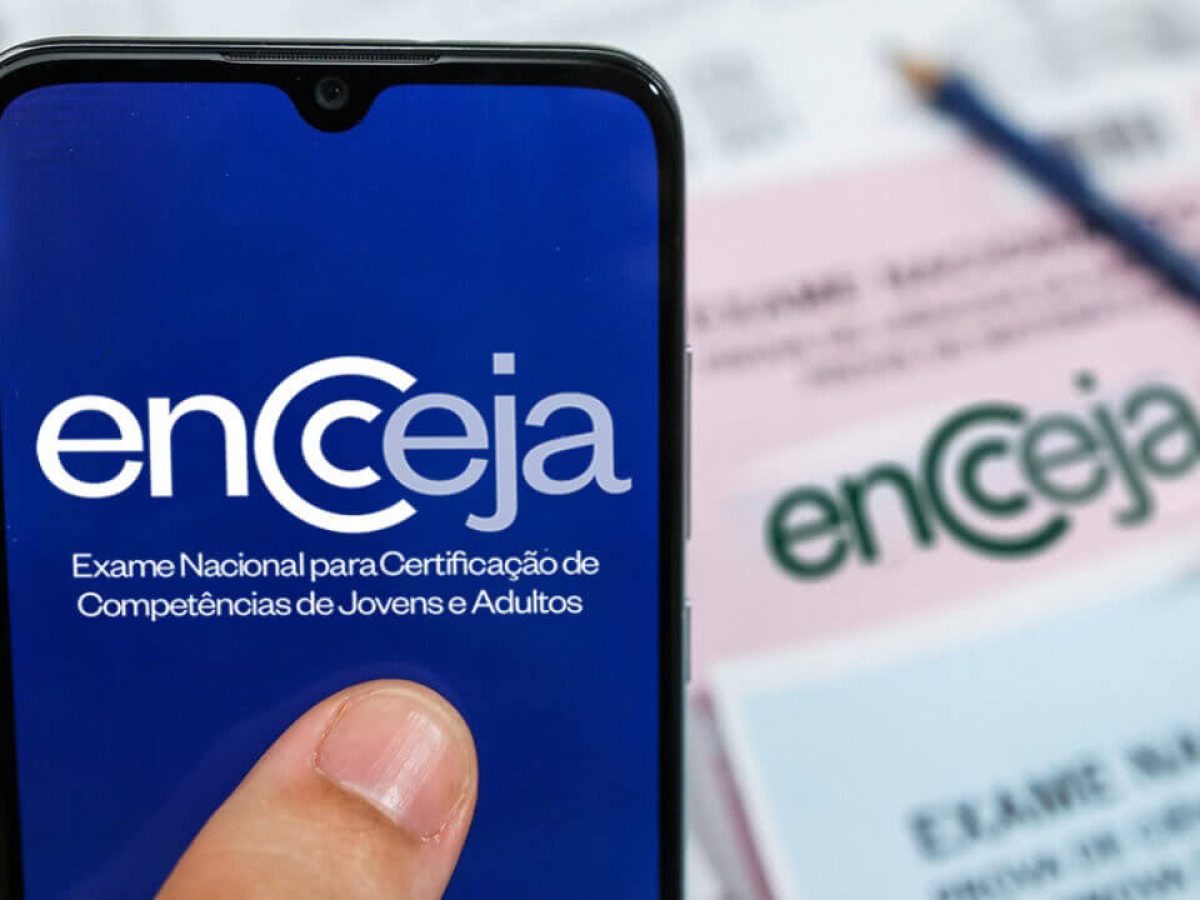 Imagem ilustrativa de dedo tocando a tela de um celular com a marca do Encceja, ao lado de cartões-resposta do exame, em referência à consulta do resultado