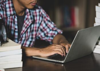 Jovem de camisa xadrez digitando em notebook rodeado de livros sobre mesa, simbolizando a consulta de cursos e instituições no e-MEC.