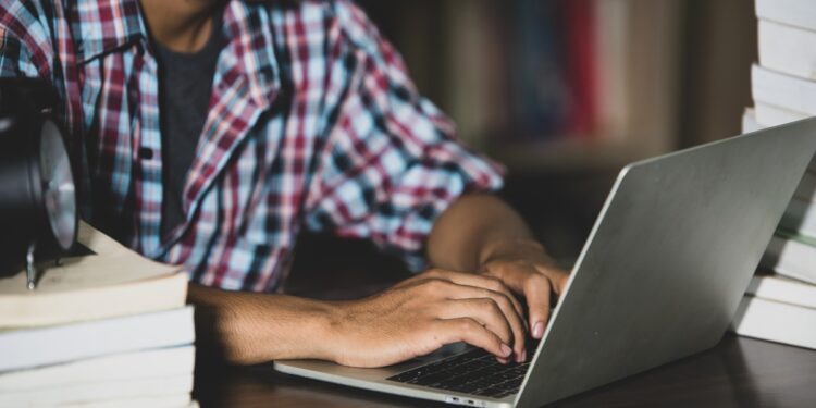 Jovem de camisa xadrez digitando em notebook rodeado de livros sobre mesa, simbolizando a consulta de cursos e instituições no e-MEC.