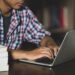 Jovem de camisa xadrez digitando em notebook rodeado de livros sobre mesa, simbolizando a consulta de cursos e instituições no e-MEC.
