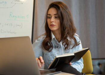 Mulher jovem estudando programação em laptop, segurando caderno e com quadro branco ao fundo, simbolizando os cursos gratuitos em IA.
