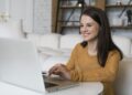 Mulher jovem sorridente usando notebook em sala aconchegante com blusa mostarda, simbolizando a importância dos cursos livres no currículo sem experiência.