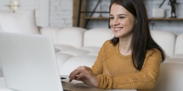 Mulher jovem sorridente usando notebook em sala aconchegante com blusa mostarda, simbolizando a importância dos cursos livres no currículo sem experiência.