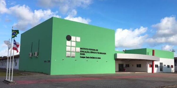 Fachada verde de um campus do Instituto Federal da Bahia, com bandeiras do Brasil e do estado hasteadas na entrada, em referência às unidades que recebem as vagas.