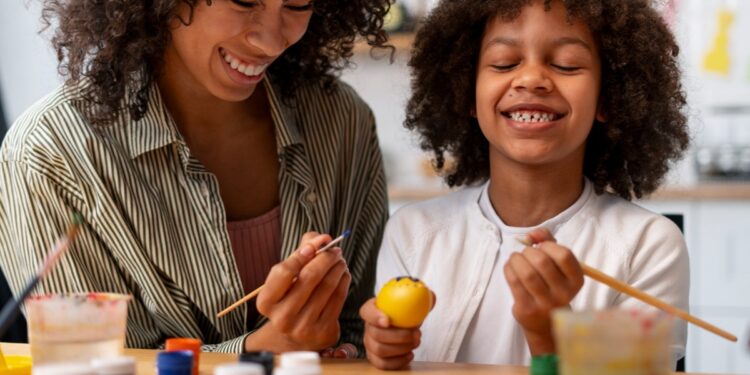 Imagem ilustrativa de educadora e estudante sorrindo enquanto pintam com pincéis e potes de tinta sobre a mesa, tema da matéria sobre as diretrizes que tornam a arte campo de conhecimento na educação básica.