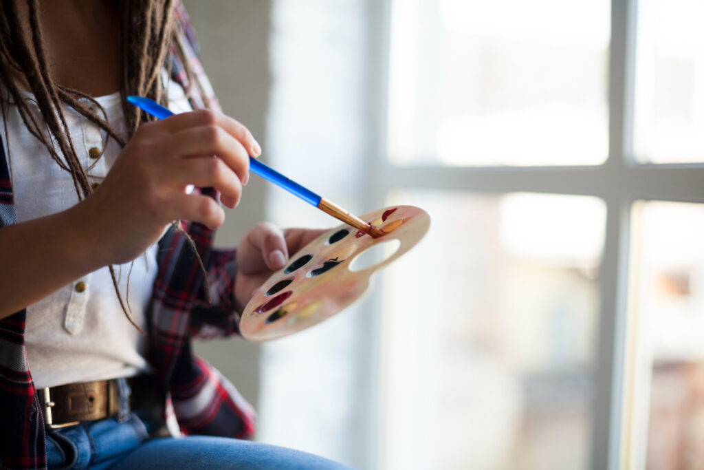 Imagem ilustrativa de estudante segurando uma paleta com tintas e um pincel azul, sentada perto de uma janela iluminada, em referência à organização da carga horária das disciplinas de arte.