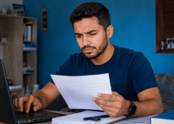 Imagem ilustrativa de candidato comparando um documento impresso com a tela do notebook, com expressão de desconfiança, cena ligada à checagem de anúncios de concurso público.