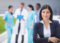 Imagem ilustrativa de gestora hospitalar de terno escuro em primeiro plano, com equipe de saúde uniformizada conversando ao fundo, representando a área administrativa da saúde.