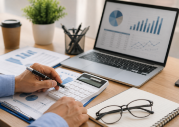 Mãos masculinas usando calculadora cercadas por gráficos, laptop com dashboard de finanças e óculos sobre caderno