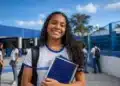Estudante com livros e mochila em frente a uma escola pública, representando o Dia do Estudante.