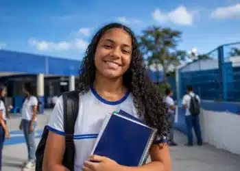 Estudante com livros e mochila em frente a uma escola pública, representando o Dia do Estudante.