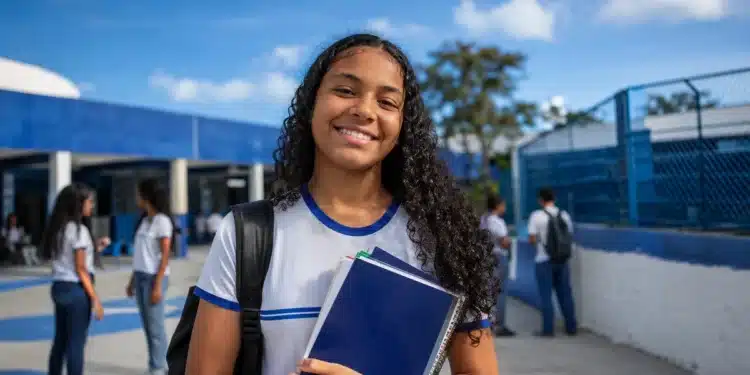 Estudante com livros e mochila em frente a uma escola pública, representando o Dia do Estudante.