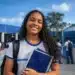 Estudante com livros e mochila em frente a uma escola pública, representando o Dia do Estudante.