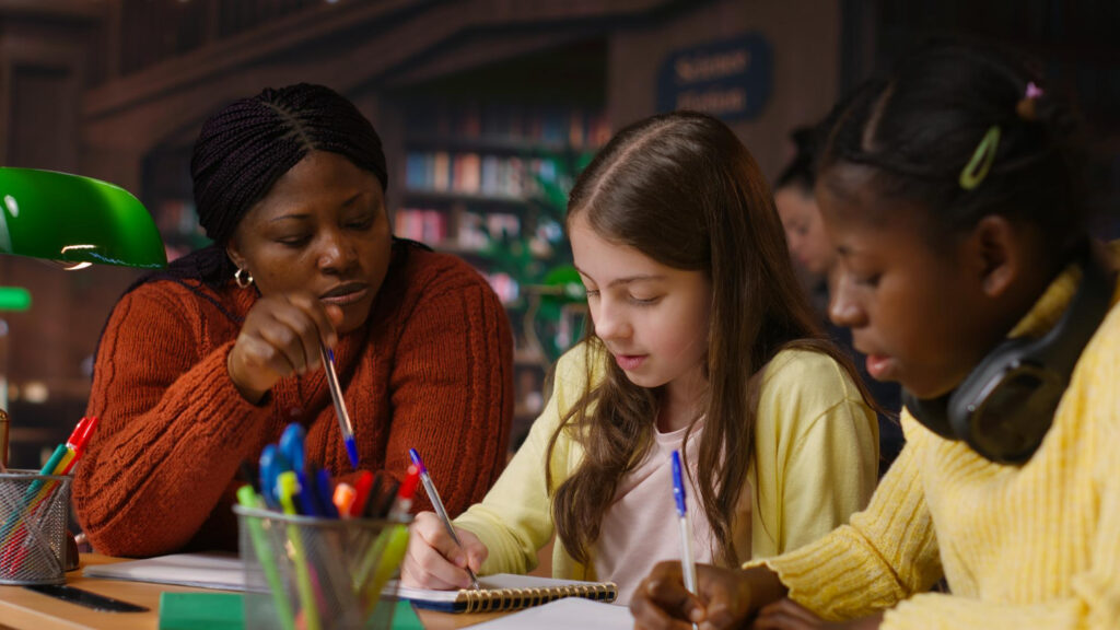 Imagem ilustrativa de professora acompanhando duas estudantes que escrevem em cadernos dentro de uma biblioteca, em referência às diferenças de desempenho escolar entre estados e regiões