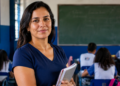 Professora posando com cadernos nos braços diante de uma turma em sala de aula de escola pública, em referência às vagas de mestrado presencial abertas pelo MEC e pela Capes