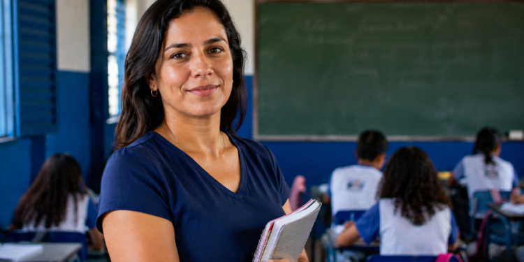 Professora posando com cadernos nos braços diante de uma turma em sala de aula de escola pública, em referência às vagas de mestrado presencial abertas pelo MEC e pela Capes