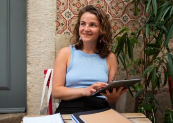 Imagem ilustrativa de mulher de regata azul-clara sentada a uma mesa externa com tablet nas mãos e cadernos à frente, tema da matéria sobre o curso gratuito de inteligência artificial para mulheres empreendedoras.