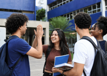 Imagem ilustrativa de grupo de estudantes com mochilas e cadernos conversando e batendo as mãos em frente à fachada azul de uma universidade, referência à ampliação da oferta de cursos de graduação no país