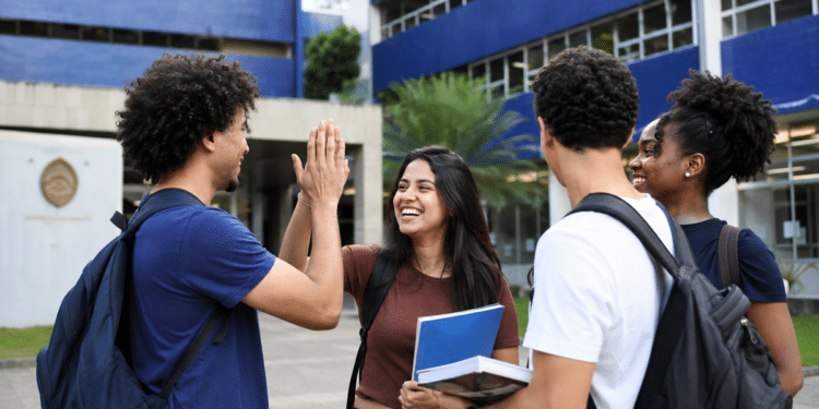 Imagem ilustrativa de grupo de estudantes com mochilas e cadernos conversando e batendo as mãos em frente à fachada azul de uma universidade, referência à ampliação da oferta de cursos de graduação no país