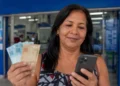 Mulher consulta o celular enquanto segura dinheiro, representando os pagamentos dos benefícios do INSS referentes a agosto e a consulta de informações pelo Meu INSS e pelo telefone 135.