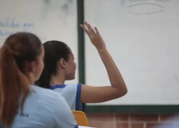 Estudante em sala de aula levanta a mão durante atividade escolar.