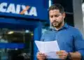 Homem segura documento em frente a uma agência da Caixa, representando o saldo do FGTS retido após pedido de demissão.