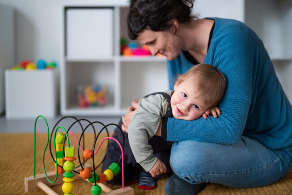 Imagem ilustrativa de profissional de blusa azul agachada no chão de uma sala de brinquedos, amparando um bebê apoiado em seu braço, em referência ao perfil exigido na função.