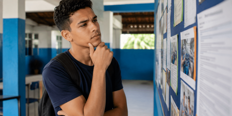 Imagem ilustrativa de jovem com mochila observando pensativo um mural de avisos em um corredor escolar, representando a decisão sobre qual curso técnico seguir