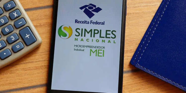 Imagem ilustrativa de celular apoiado em mesa de madeira exibindo as marcas da Receita Federal, do Simples Nacional e do MEI, ao lado de calculadora e agenda, em referência às mudanças no regime a partir de 2027