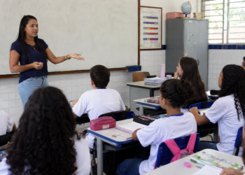 Professora da educação básica explica conteúdo para alunos em sala de aula de escola pública.
