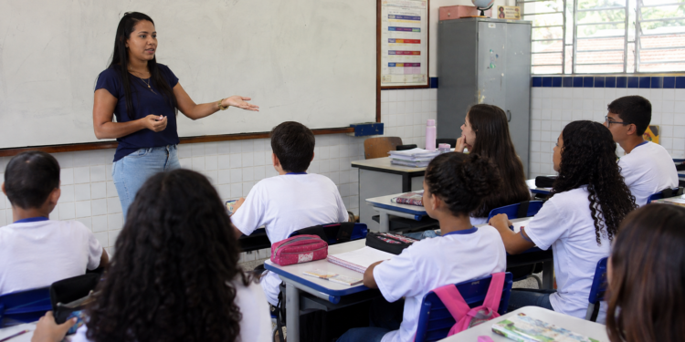 Professora da educação básica explica conteúdo para alunos em sala de aula de escola pública.