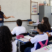 Professora da educação básica explica conteúdo para alunos em sala de aula de escola pública.