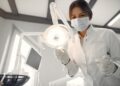 Imagem ilustrativa de profissional de jaleco branco posicionando o refletor odontológico com instrumentos nas mãos dentro do consultório, em referência aos termos dentista, odontólogo e cirurgião-dentista