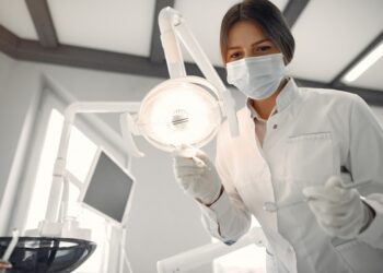 Imagem ilustrativa de profissional de jaleco branco posicionando o refletor odontológico com instrumentos nas mãos dentro do consultório, em referência aos termos dentista, odontólogo e cirurgião-dentista