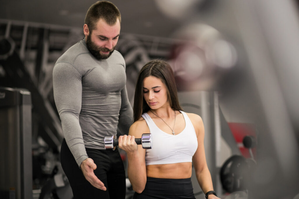 Imagem ilustrativa de personal trainer orientando aluna durante exercício com halter na academia, em referência às etapas para começar a atuar na profissão