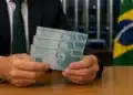 Homem de terno segura três cédulas de R$ 100 em ambiente institucional com bandeira do Brasil ao fundo