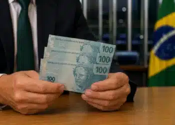 Homem de terno segura três cédulas de R$ 100 em ambiente institucional com bandeira do Brasil ao fundo