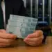 Homem de terno segura três cédulas de R$ 100 em ambiente institucional com bandeira do Brasil ao fundo