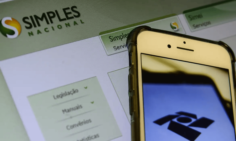 Imagem ilustrativa de celular com a marca da Receita Federal na tela diante do portal do Simples Nacional aberto no computador, em referência ao prazo de adesão ao regime