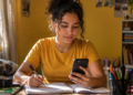 Imagem ilustrativa de estudante fazendo anotações em caderno enquanto consulta o celular no quarto, em referência ao simulado gratuito do Enem disponível no Gemini