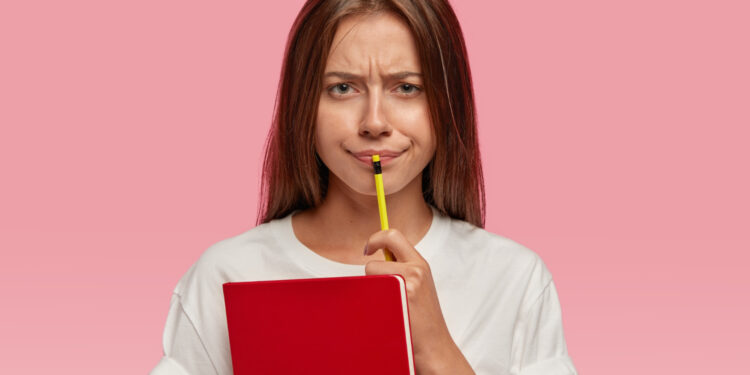 Imagem ilustrativa de jovem de camiseta branca segurando um caderno vermelho e um lápis, com expressão de dúvida, em referência ao emprego de por que em perguntas.