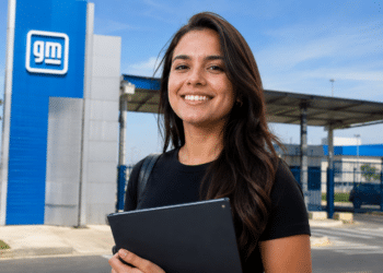 Imagem ilustrativa de jovem profissional sorridente segurando um tablet diante da entrada de uma unidade da General Motors.