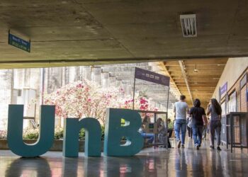 Letreiro tridimensional com a sigla UnB instalado em corredor coberto do campus da Universidade de Brasília, com estudantes caminhando ao fundo.