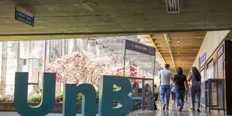 Letreiro tridimensional com a sigla UnB instalado em corredor coberto do campus da Universidade de Brasília, com estudantes caminhando ao fundo.