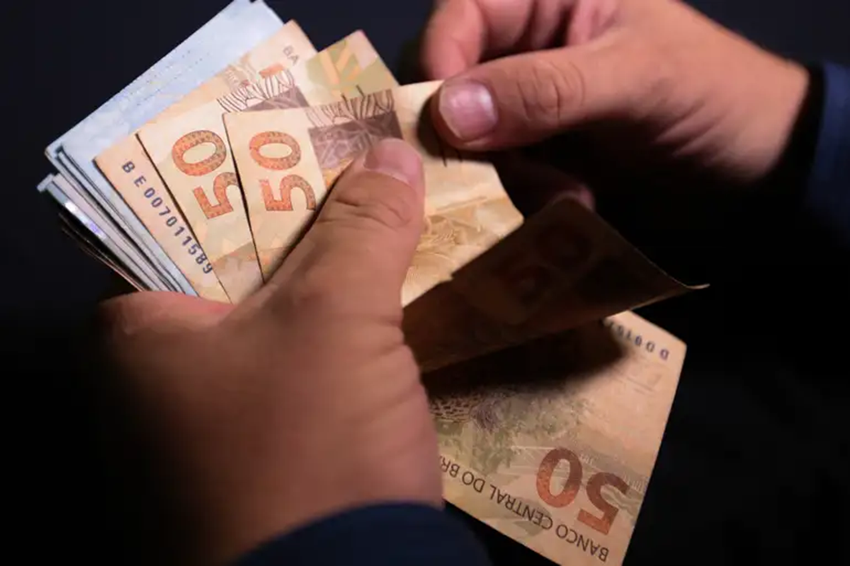 Mãos masculinas segurando e contando cédulas de cinquenta reais do Banco Central do Brasil, simbolizando o pagamento do 13º salário do INSS.
