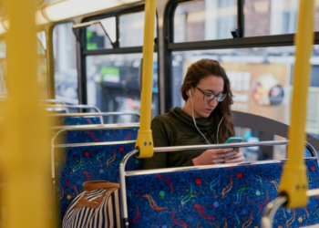 Jovem usando celular com fones em ônibus vazio durante o dia.