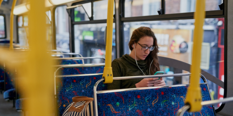 Jovem usando celular com fones em ônibus vazio durante o dia.