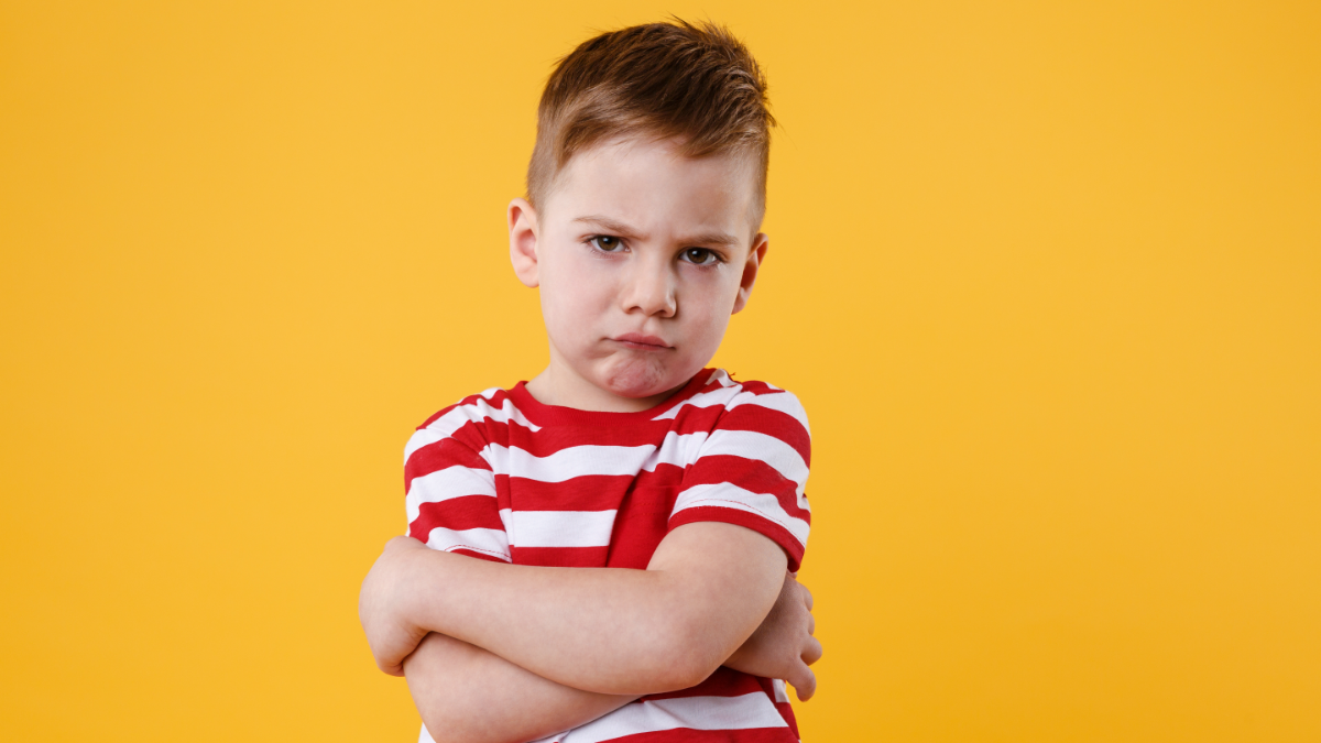 Menino com expressão emburrada de braços cruzados em fundo amarelo.